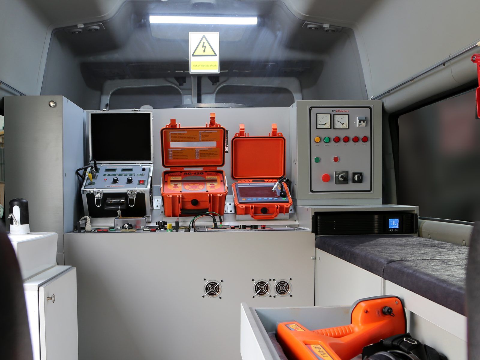Inside the van: an instrument bench with an aluminium-cased instrument and two orange instrument cases, a control panel with meters and lamps, a UPS below it, and a pull-out drawer holding a hand-held receiver and headphones.